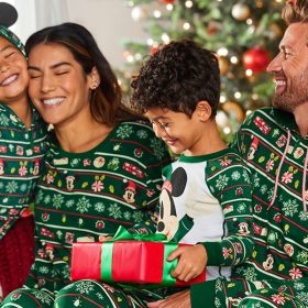 A family is sitting on a couch in matching green holiday pajamas with a Christmas tree in the background feeling like Christmas morning. The pajamas have multiple holiday and Disney icons arranged on rows throughout. The mom is hugging the daughter as the dad looks at them with a smile. The little boy sitting between his parents has a present wrapped in red paper with a green bow in his lap that he’s ready to open it.