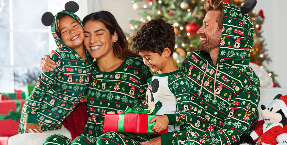 A family is sitting on a couch in matching green holiday pajamas with a Christmas tree in the background feeling like Christmas morning. The pajamas have multiple holiday and Disney icons arranged on rows throughout. The mom is hugging the daughter as the dad looks at them with a smile. The little boy sitting between his parents has a present wrapped in red paper with a green bow in his lap that he’s ready to open it.