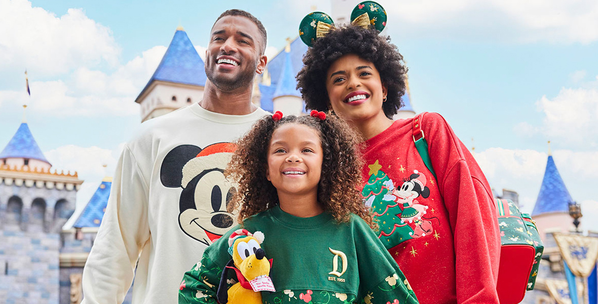 A man, woman, and young girl are standing in front of a Disney castle wearing holiday crewnecks from DisneyStore.com. They man’s crewneck is a light cream color with a large image of Mickey wearing a Santa hat. The woman is wearing a red holiday crewneck with an image of Minnie decorating a Christmas tree. The young girl is holding a plushie Pluto and is wearing a green Disneyland holiday crewneck with a large D on the front.