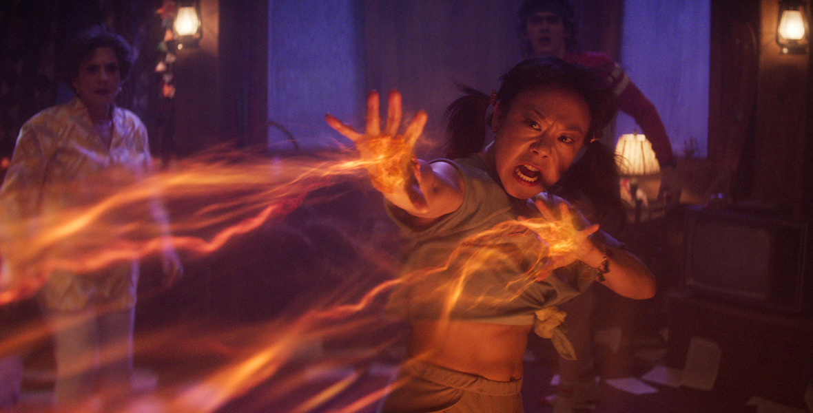 In a scene from Agatha All Along, Alice Wu-Gulliver (played by Ali Ahn) extends her hands, releasing orange neon beams of magic. Her face reflects the effort of conjuring the magic. In the background, Lilia Calderu (played by Patti LuPone) and Teen (played by Joe Locke) watch in shock.