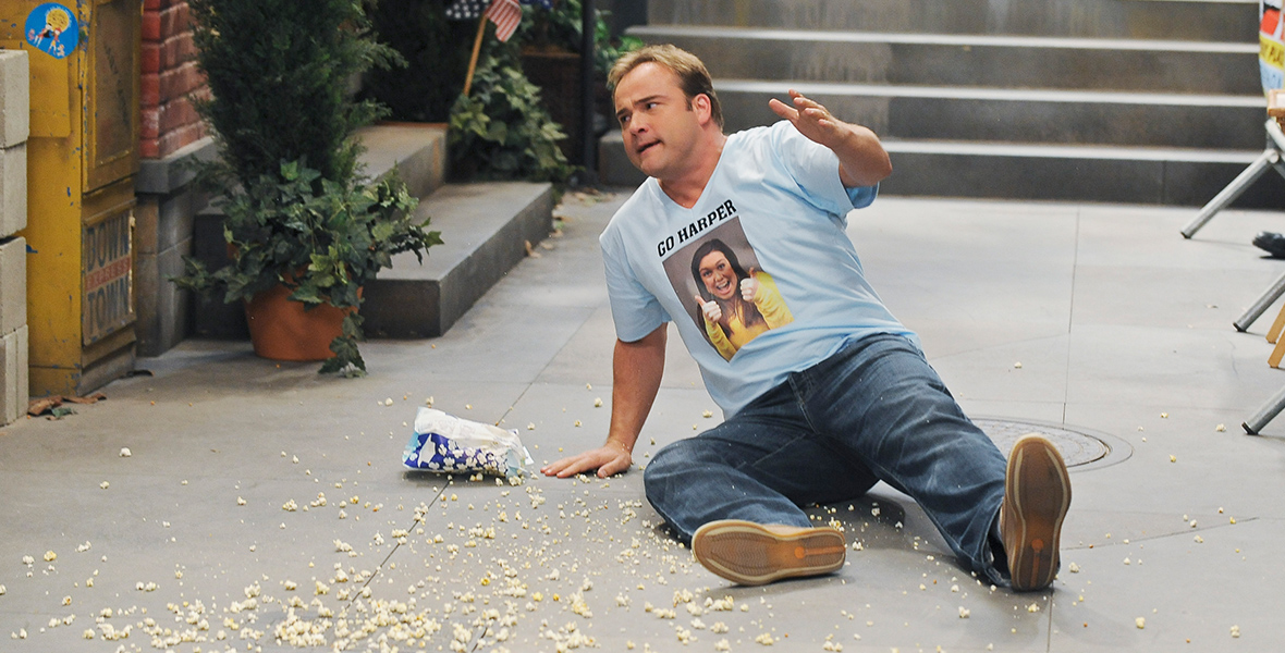 In a scene from Disney Channel’s Wizards of Waverly Place, Jerry Russo (David DeLuise) is sitting on the pavement, looking frustrated. Next to him is a spilled bag of popcorn. He is wearing a light blue graphic t-shirt with a photo of Harper Finkle (Jennifer Stone) and the words 'GO HARPER,' along with blue jeans and brown loafers. In the background are stairs and the lower half of a building’s entrances.