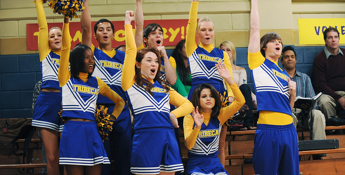 In a scene from Disney Channel’s Wizards of Waverly Place, Alex Russo (Selena Gomez) sits on the wooden bleachers of an indoor gym while her fellow cheerleaders stand and perform a dance; Alex, is seated with her hands slightly raised. The team’s uniform is blue, yellow, and white, with 'TRIBERA' across the front. The background shows other people sitting on the bleachers, with a yellow and blue painted stone wall behind them.