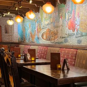 An image of the La Cava del Tequila restaurant interior. Rustic and cozy with circular lamps hang down from the ceiling and wooden chairs are pushed into wooden candlelit tables. The Mexican tequila bar features a unique handmade mural over a brick wall, depicting the process of the agave spirits distillation—from the harvesting and the cooking, to the fermentation, distillation, and aging in the big brown barrels.