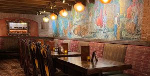 An image of the La Cava del Tequila restaurant interior. Rustic and cozy with circular lamps hang down from the ceiling and wooden chairs are pushed into wooden candlelit tables. The Mexican tequila bar features a unique handmade mural over a brick wall, depicting the process of the agave spirits distillation—from the harvesting and the cooking, to the fermentation, distillation, and aging in the big brown barrels.