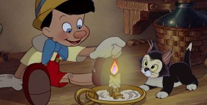 In a scene from Pinocchio (1940), Pinocchio (voiced by Dickie Jones), a living wooden puppet, wears a pointy yellow hat, a yellow shirt, red overalls, a blue bow tie, and maroon shoes. He reaches out with his left finger to touch a lit candle. On the right side of the frame, a small black-and-white cat looks alarmed at Pinocchio's action, with a wooden shelf in the background. The scene is dimly lit.