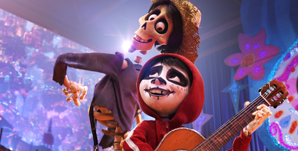 In a scene from Coco (2017), Héctor (voiced by Gael García Bernal), a skeletal spirit, wears a straw hat and a tattered long-sleeve shirt. Miguel (voiced by Anthony Gonzalez) wears a red zip-up jacket with a hoodie and holds a guitar, looking up at Héctor. Miguel's face is painted white with black around his eyes, nose, and mouth. The two are pictured dancing on a stage, with a colorful backdrop in the background.