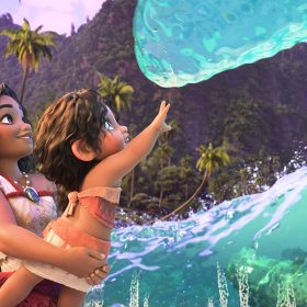 In an image from Walt Disney Animation Studios’ Moana 2, Moana (voiced by Auli‘i Cravalho) is holding her little sister Simea (voiced by Khaleesi Lambert-Tsuda) up to touch a wave; they are standing on a Motunui beach, surrounded by the water, which is acting friendly to the pair as it has to Moana in the past. There are lush green mountains and palm trees seen in the background.