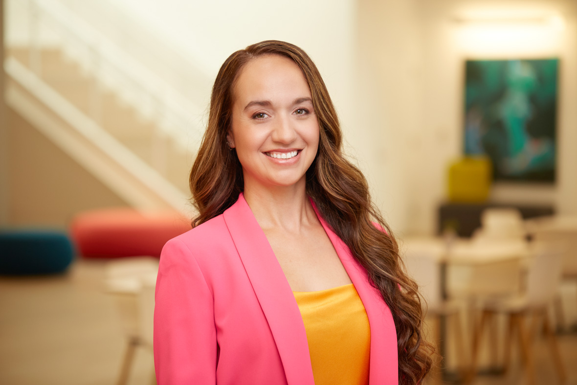 A headshot of The Art of Moana 2 author Kalikolehua Hurley; she is wearing a pink blazer and a yellow silk top, and has long light brown hair. She smiles at the camera.