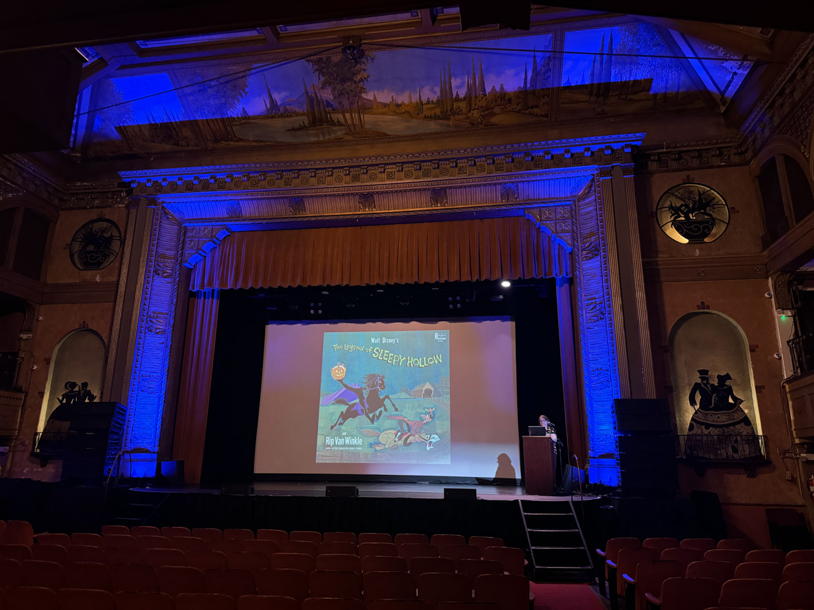 Becky Cline, Director of the Walt Disney Archives, giving a presentation on stage inside Tarrytown Music Hall. The screen beside her features a graphic depicting art from Walt Disney’s The Legend of Sleepy Hollow.