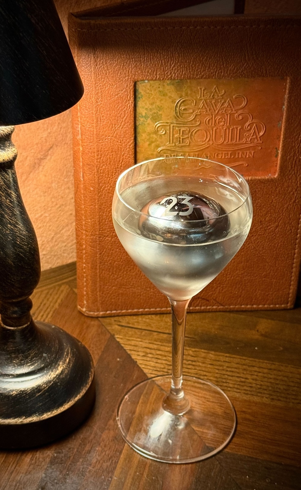 The Aztec Eagle cocktail is presented in tall clear cocktail glass and sits on a wooden table under the glow of a table lamp, in front of a La Cava del Tequila menu. Floating in the cocktail is a frozen metal sphere, personalized and engraved with a “23” representing the cocktail being exclusive to D23 members. The menu is made of leather and sports a soft orange color. A rectangular cutout in the top middle of the menu features the La Cava del Tequila logo; it almost looks etched into the leather material itself.