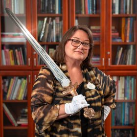 Walt Disney Archives director Becky Cline, wearing a patterned shawl and white gloves, poses confidently while holding the “Excalibur,” a decorative prop sword from the 2004 film King Arthur, with shelves filled with books in the background.