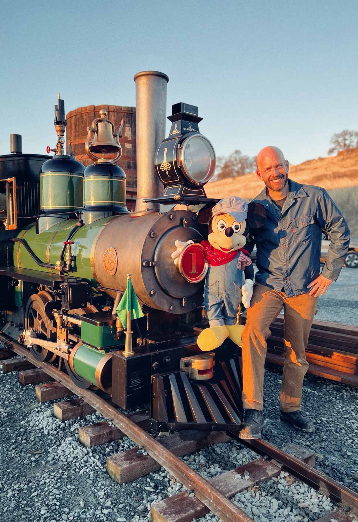 A green and black half-scale steam engine train with gold accents is parked on a railroad, with Bret Iwan standing in front, posing for the photo. He holds a large Mickey Mouse doll, with Mickey’s arm resting on the “1” sign displayed on the front of the train. Bret is dressed in a blue shirt, brown pants, and dark brown loafers. The background features a clear sky and golden, wheat-colored hills.