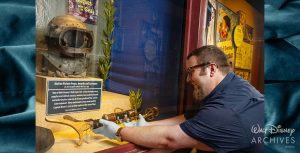 Kevin Kern, a member of the Walt Disney Archives staff, wearing a short-sleeved blue shirt, a pair of glasses, a wristwatch, and white archival gloves, carefully positions a brown and gold-colored undersea rifle prop from the 1954 film 20,000 Leagues Under the Sea in an open display case. Also displayed are a rusted dive helmet, prop sea foliage, and a descriptive text panel of the items on display.