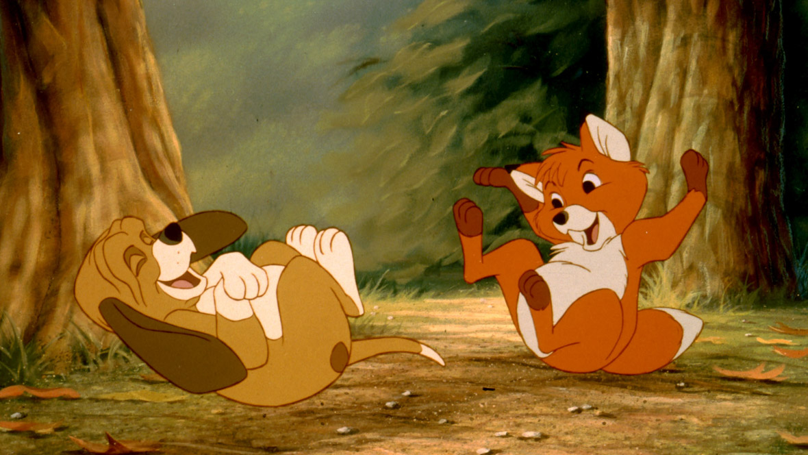 In a scene from The Fox and the Hound, Copper (voiced by Corey Feldman), a hound dog, and Tod (voiced by Keith Coogan), a fox, are playfully running and laughing in the woods. The background features two trees and scattered dry leaves on the forest floor.