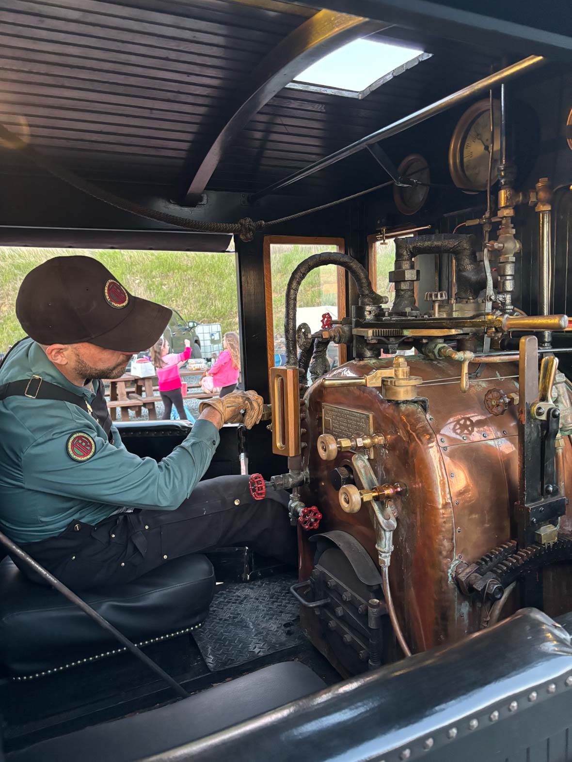 Bret Iwan is inside a train, wearing a blue shirt, dark blue overalls, a brown cap, and light brown gloves. He is seated on a black leather seat framed by dark brown wooden accents, working on the train's engine.