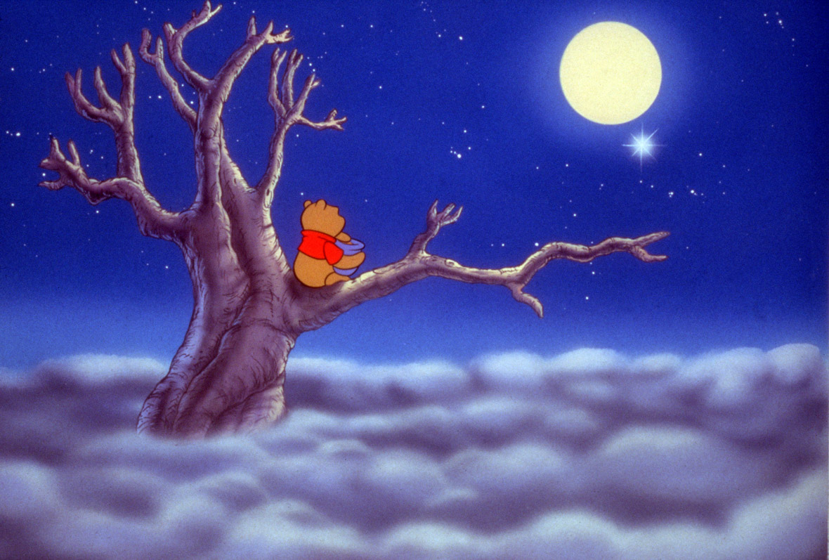 In a scene from Pooh’s Grand Adventure, Winnie the Pooh (voiced by Jim Cummings) sits perched on the branch of a large tree, surrounded by a dreamy expanse of soft, fluffy clouds. The nighttime sky is deep blue with stars and a full, glowing moon. Pooh wears his signature cropped, short-sleeved red top; cradles a honey pot in his arms; and looks at the moon and stars.