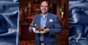 Steven Vagnini, a member of the Walt Disney Archives staff, wearing a blue sports coat, blue-checkered long-sleeved shirt, and white archival gloves in a warm museum-like setting, smiles at the camera while holding a small handcar toy that is pulled by a miniature Mickey Mouse and Minnie Mouse.