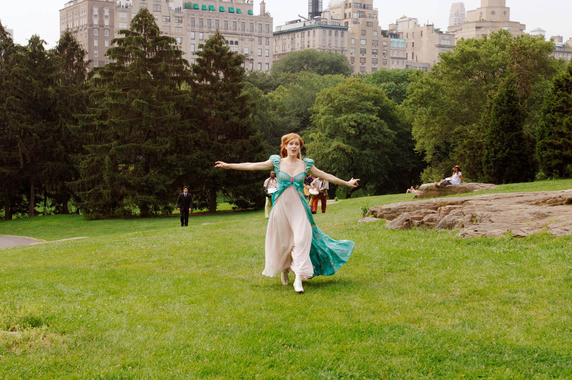 In a scene from Enchanted, Giselle (Amy Adams) runs through Central Park with her arms outstretched. She wears a short-sleeved blue dress with a sweetheart neckline and colorful accents, opening at the front to reveal a white, crystal-embellished underskirt. In the background, people play the cajón drum, while Robert Philip (Disney Legend Patrick Dempsey) follows Giselle. Tall trees frame the park, with buildings rising in the distance. 