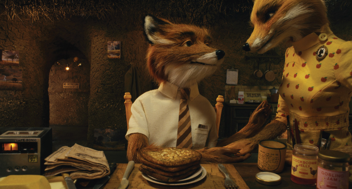 In a scene from Fantastic Mr. Fox, Mr. Fox (voiced by George Clooney), an anthropomorphic fox, sits at a table and looks at Mrs. Fox (voiced by Meryl Streep) as she sets down a heart-shaped dish. Mr. Fox is dressed in a short-sleeved white shirt with a striped tie, while Mrs. Fox wears a yellow wrap dress adorned with small red apple details. The background reveals the cozy interior of the Fox family's dining and kitchen area.