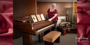 Laura Sanchez, a member of the Walt Disney Archives staff, wearing a red-and-black patterned blouse and white archival gloves, has her hands placed on Walt Disney’s piano, which has been opened to reveal the keyboard. Displayed on top of the piano are a Mousecar award statue (in the shape of Mickey Mouse), a row of sheet music, and a small statue of the Little Mermaid from Europe’s Tivoli Gardens. The warm office setting, featuring wood paneling, also includes an upholstered piano bench, a glass table, and a light-colored couch.