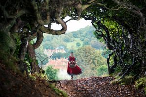 In a scene from&nbsp;Into the Woods, Little Red Riding Hood (Lilla Crawford) skips along a path covered in fallen leaves. Encircling the frame are twisted branches intertwined with grass and foliage. In the background, pointed-roof houses and dense trees are visible. Little Red wears a short-sleeved blue dress beneath a red cloak and carries a woven basket.