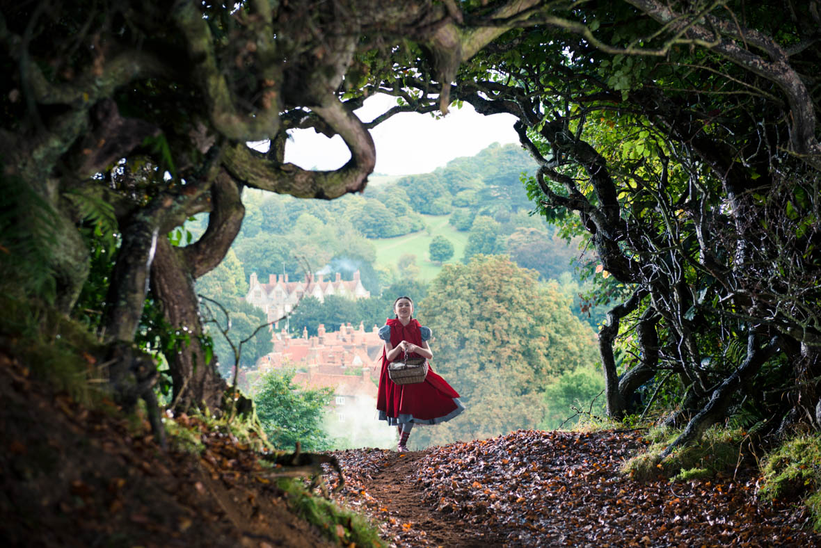 In a scene from Into the Woods, Little Red Riding Hood (Lilla Crawford) skips along a path covered in fallen leaves. Encircling the frame are twisted branches intertwined with grass and foliage. In the background, pointed-roof houses and dense trees are visible. Little Red wears a short-sleeved blue dress beneath a red cloak and carries a woven basket.