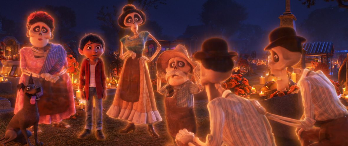 In a scene from Coco, Miguel (voiced by Anthony Gonzalez), dressed in a red hoodie over a white shirt and blue jeans, stands wide-eyed and surprised in the cemetery, surrounded by glowing skeletal ancestors. Next to Miguel stands Dante, a black dog, with his tongue out and ears perked up. The background features a cemetery bedecked in glowing candles and flowers.