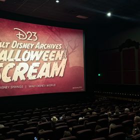 Guests in the Disney Springs AMC Theatre with the “D23 Walt Disney Archives Halloween Scream” logo on the silver screen. Image of yellow, large and bold font that reads “D23 Walt Disney Archives Halloween Scream Disney Springs, Walt Disney World” against a copper background with bare, scary tree silhouettes. There are also branches growing from the “S” and the “M” in the word “scream”