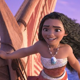 In an image from Walt Disney Animation Studios’ Moana 2, Moana (voiced by Auli‘i Cravalho) is seen on her canoe in the middle of the ocean, steering it and looking excitedly off camera to the left. Her hair is windswept, and the sky and seas are calm.