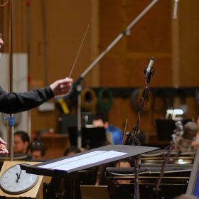 In an image from the Music by John Williams documentary, John Williams is conducting an orchestra. He wears a black mock turtleneck sweater and black pants, holding a baton as he looks downward at a large music stand elevated on a black podium. The blurred background features the wooden interior of the room, with microphones and orchestra members playing instruments, including violins.