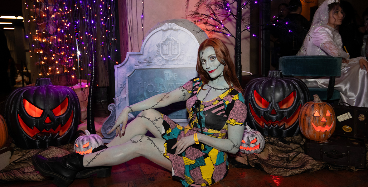 A guest dressed as Sally from Tim Burton’s The Nightmare Before Christmas strikes a pose in front of vibrant Halloween decor, including glowing jack-o’-lanterns and a spooky sign for The Hollywood Tower Hotel. The scene is illuminated by enchanting purple and orange lights, creating a magical and atmospheric backdrop.