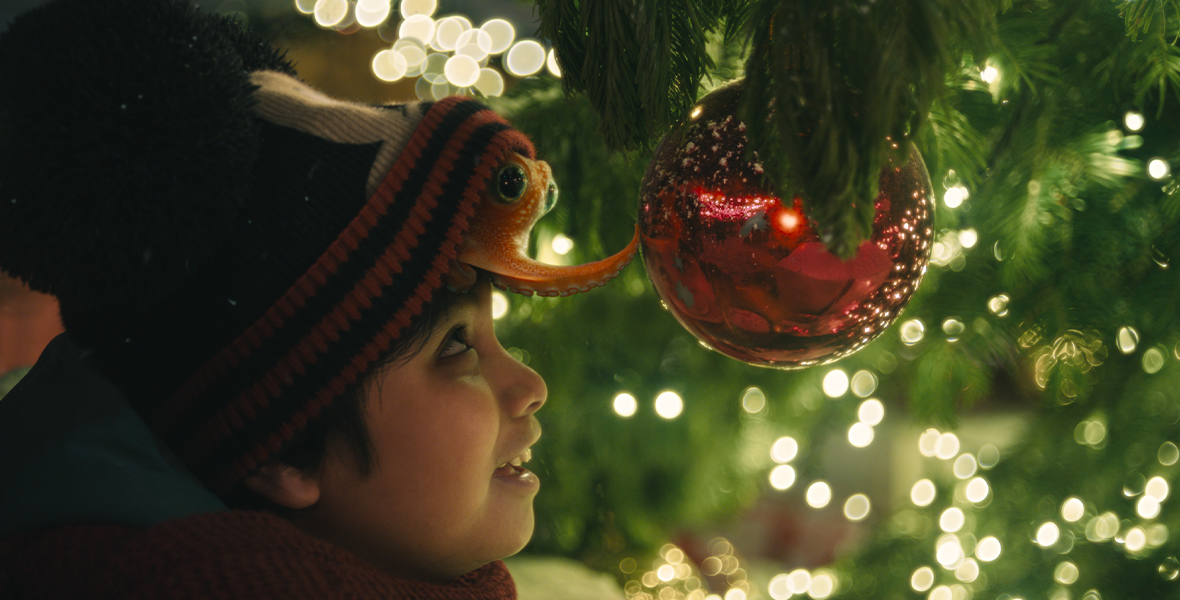 An orange octopus, sitting atop a young boy's head, peeks out from underneath a Mickey Mouse beanie. It reaches out and notices its reflection in a red Christmas ornament.