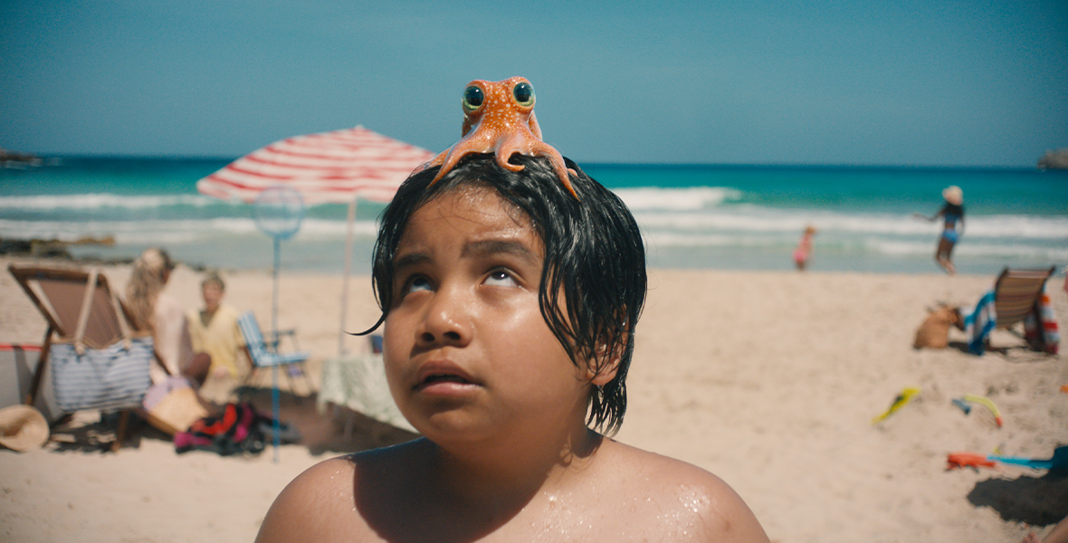 An orange octopus sits atop a young boy's head. It's a crowded day at the beach.