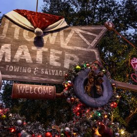 In a photo from Mater’s Jingle Jamboree at Disney California Adventure Park, a sign reading "Tow Mater" is adorned with a Santa Claus hat and festively wrapped garlands. The background showcases an afternoon sky with trees.