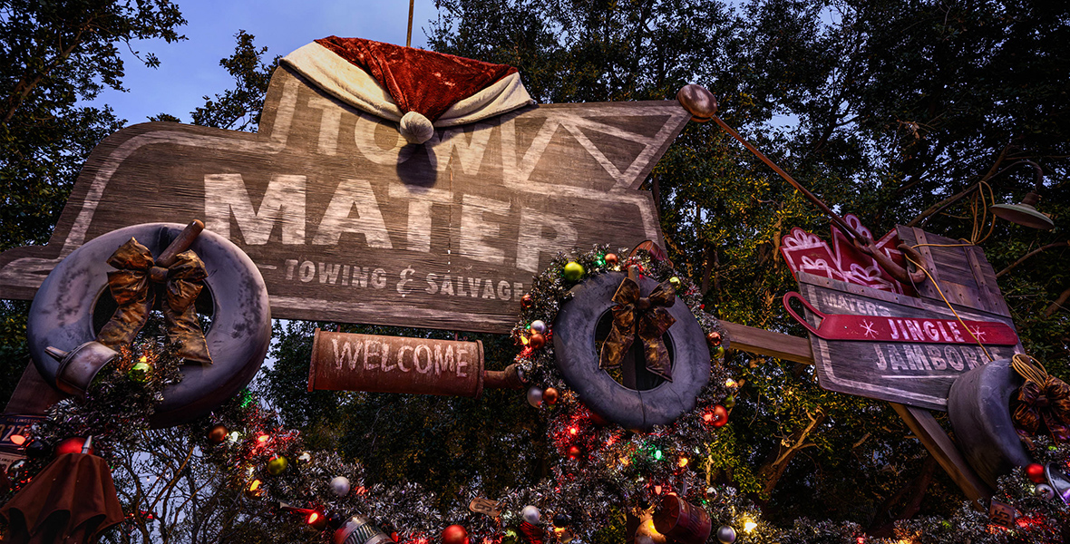 In a photo from Mater’s Jingle Jamboree at Disney California Adventure Park, a sign reading "Tow Mater" is adorned with a Santa Claus hat and festively wrapped garlands. The background showcases an afternoon sky with trees.