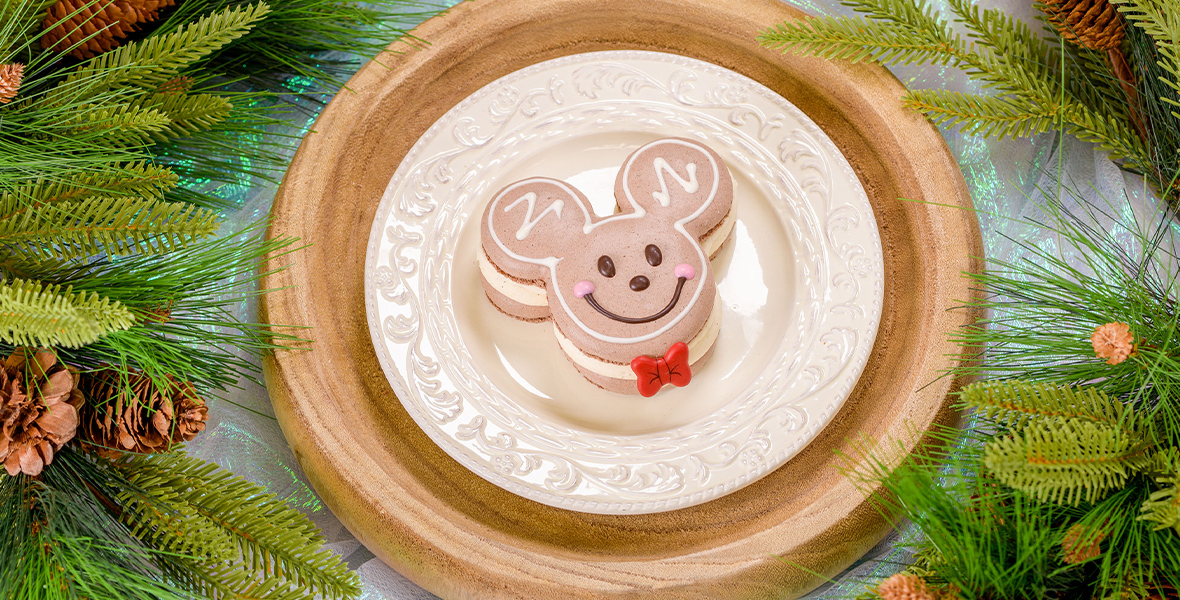 A photo featuring a plate with a holiday-themed, milk-chocolate macaron in the shape of Mickey Mouse’s head, smiling. Pinecones and pine tree leaves are arranged around the sides.