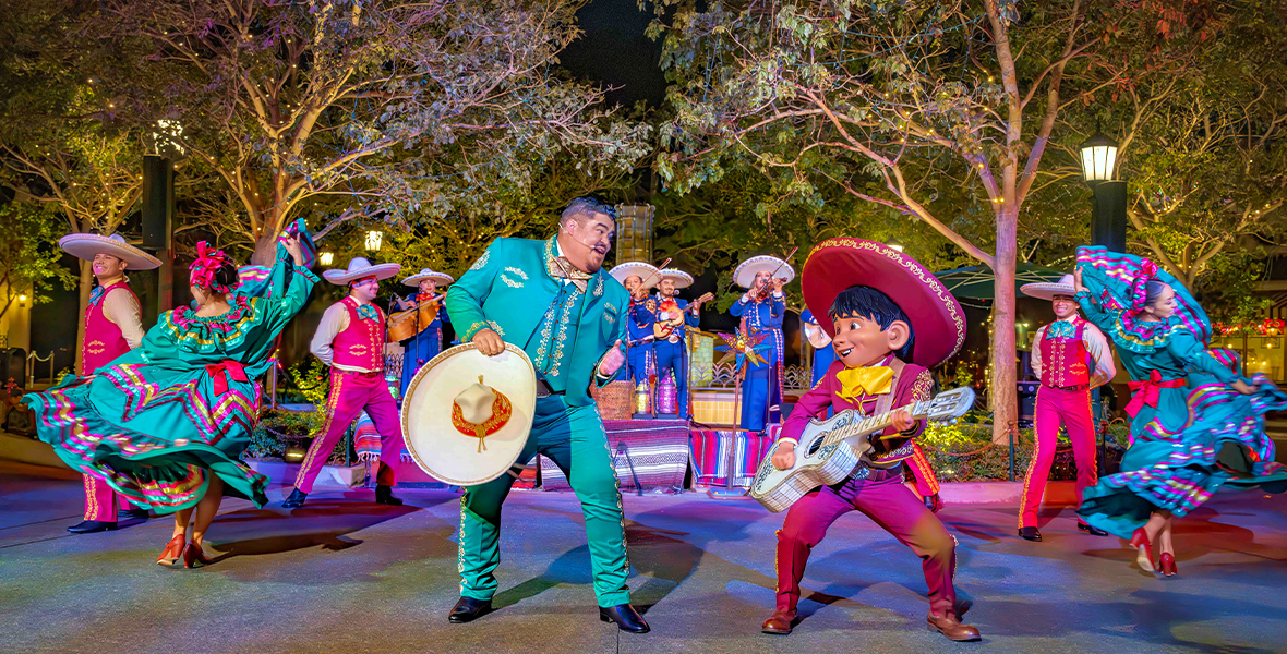 In a photo from the A Musical Christmas with Mariachi Alegría de Disneyland & Miguel!performance at Disney California Adventure, the mariachi group Mariachi Alegría de Disneyland is joined by Miguel from Pixar Animation Studios’ Coco. They wear traditional mariachi attire in red and green, while neon blue and purple lights illuminate the performance. The background features trees and a dark sky.