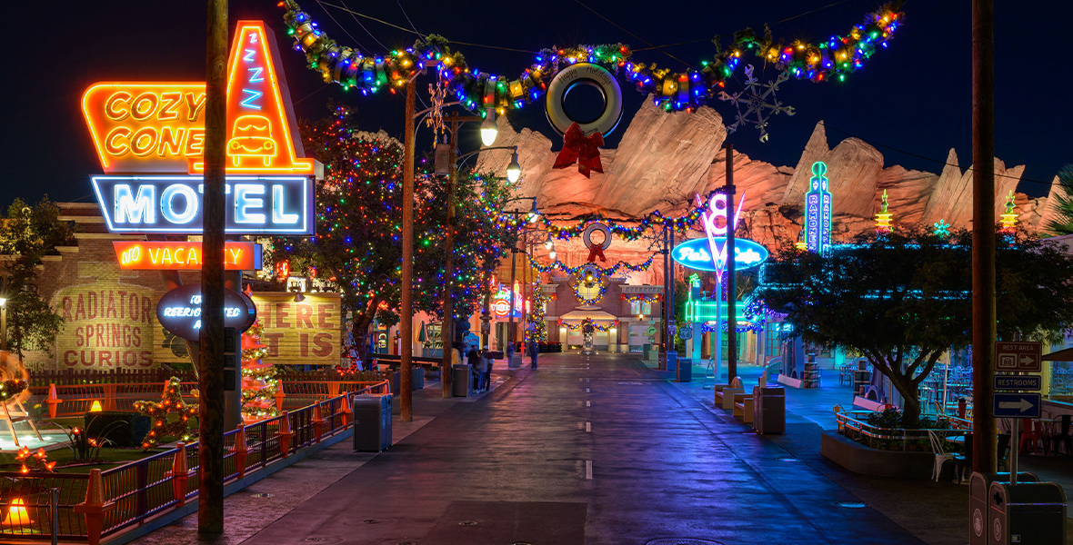 In a photo of Cars Land at Disney California Adventure, a garland decorated with Christmas lights and a wreath at its center hangs across the entrance. The background features a neon-lit sign that reads “MOTEL,” along with a lit Christmas tree and the mountain peaks of the Radiator Springs Racers ride.