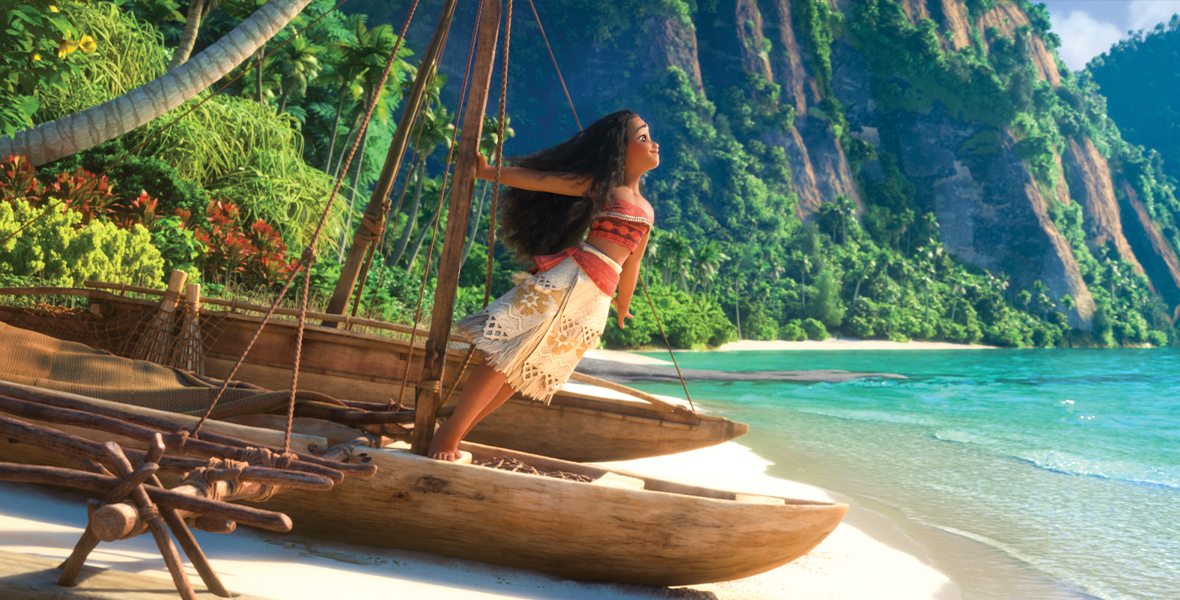 In an image from Moana, Moana (voiced by Auli’i Cravalho) stands confidently at the stern of a wooden canoe, gripping the central mast with her right hand as she leans forward. She wears a red crop top with intricate patterns paired with a white wrap skirt accented by dark yellow details. The background showcases lush green mountains, a shoreline, and sparkling crystal-clear waters.