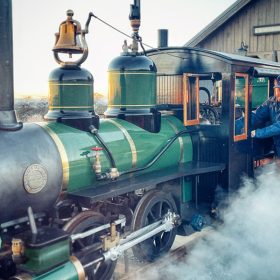 A green and black half-scale steam engine train with gold accents is parked on a railroad, emitting smoke across the tracks. Bret Iwan stands next to the train’s control panel, wearing a blue and white engineer’s cap, a blue jacket, and brown pants. A wooden house and a clear sky are visible in the background.