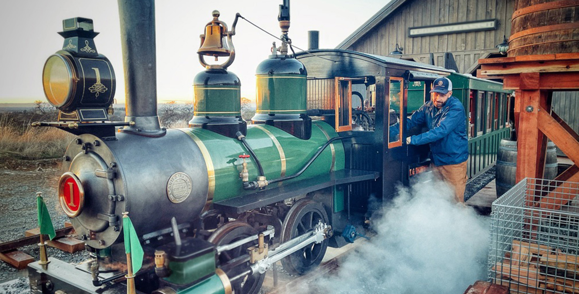 A green and black half-scale steam engine train with gold accents is parked on a railroad, emitting smoke across the tracks. Bret Iwan stands next to the train’s control panel, wearing a blue and white engineer’s cap, a blue jacket, and brown pants. A wooden house and a clear sky are visible in the background.