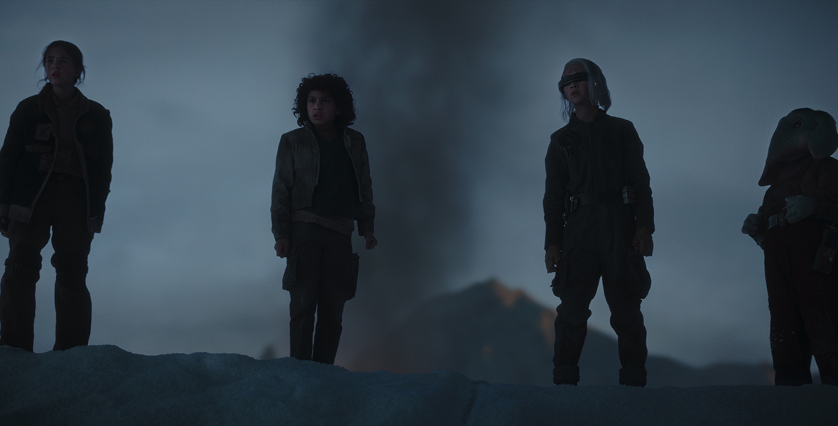 In a scene from Star Wars: Skeleton Crew, Fern, played by Ryan Kiera Armstrong, Wim played by Ravi Cabot-Conyers, KB, played by Kyriana Kratter, and Neel, played by Robert Timothy Smith, stand outside at dusk. A volcano with molten lava is in the distance.