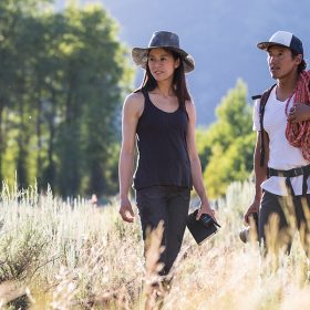 Documentary filmmakers Chai Vasarhelyi and Jimmy Chin on location during the filming of National Geographic’s Free Solo. Vasarhelyi, on the left, is wearing a black tank top, pants, and hat; Chin, on the right, is wearing a white T-shirt and dark pants and a baseball cap, and has a large bundle of rope over one shoulder. The pair is standing in a field of tall grass; green hills can be seen behind them.