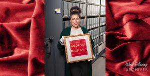 Jessica Amezcua, a member of the Walt Disney Archives staff, wearing a white blouse, green sweater, and white archival gloves, holds a gold-colored frame containing a piece of red signage that features gold-colored words reading &ldquo;ARCHIVES &ndash; ENTER RM 3.&rdquo; Behind her is an archival rolling shelf, open to a row of shelving storing dozens of gray archival boxes across a series of columns.