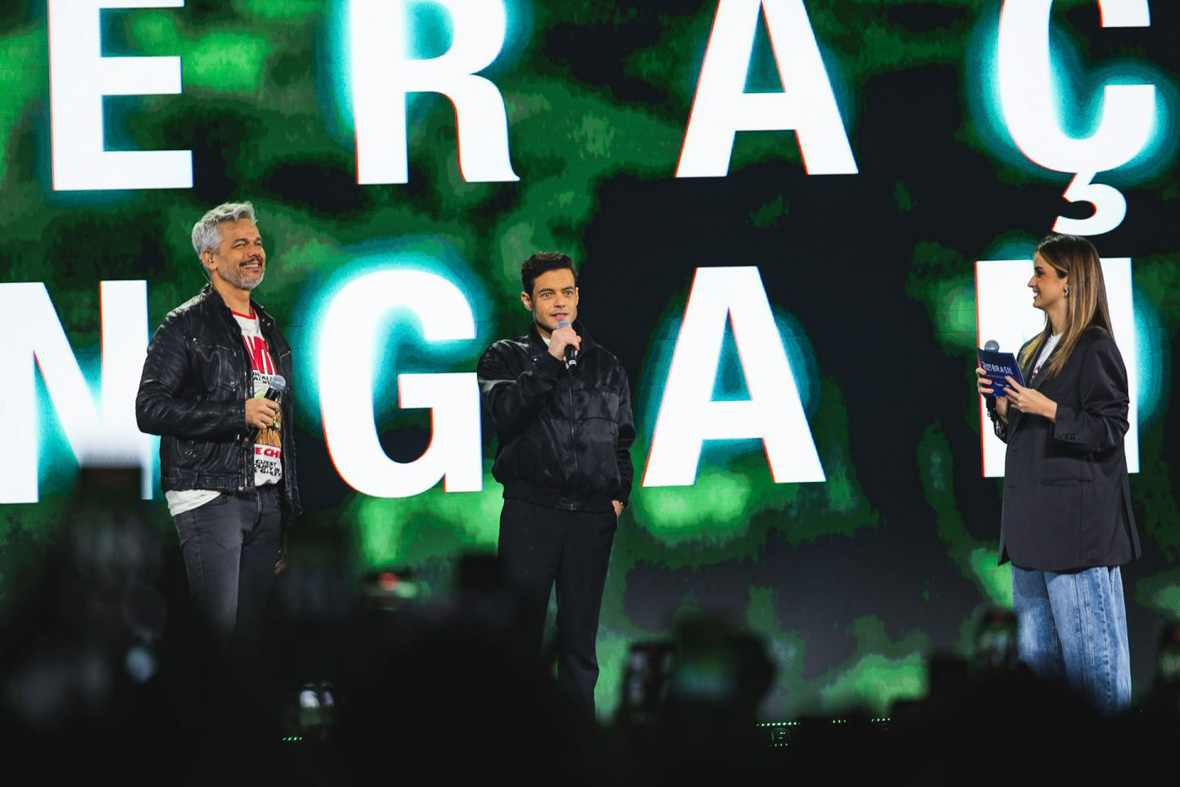 Otaviano Costa, left, Rami Malek, center, and Carol Moreira, right, speak onstage during Day 2 of the D23 Brazil – A Disney Experience at Transamerica Expo Center on November 9, 2024, in São Paulo, Brazil. Behind them is a huge video display depicting white letters against a green and black background, but the words can’t be made out.