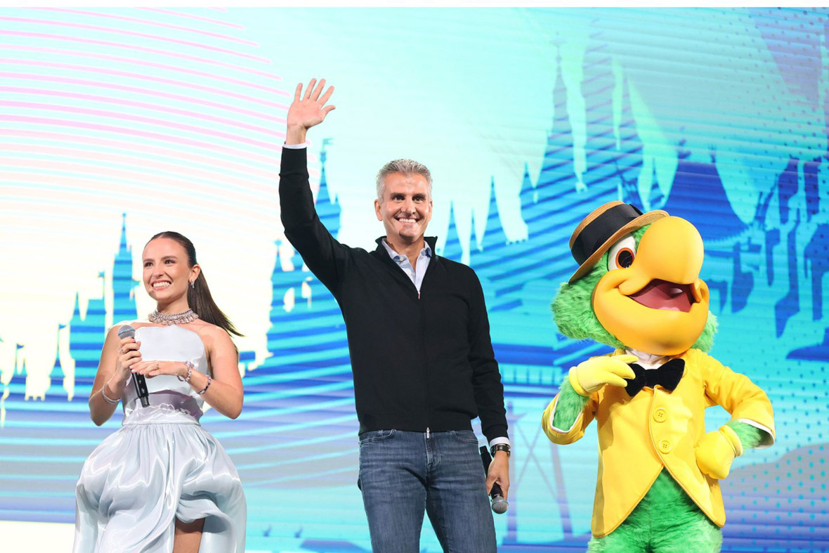 Larissa Manoela (left) and Josh D’Amaro (center), Chairman, Disney Experiences, wave to the audience during Day 2 of the D23 Brazil – A Disney Experience in São Paulo, Brazil. To D’Amaro’s right is the character José Carioca, a green parrot in a yellow suit jacket with a hat and black tie. Behind them is a video screen displaying graphic representations of the castles from Disney Parks.