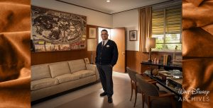 Edward Ovalle, a member of the Walt Disney Archives staff, wearing a tie and black cardigan, stands confidently in Walt Disney&rsquo;s working office, which is decorated with a large map of a Disneyland. Behind him, a beige couch and a lamp provide a cozy atmosphere, while Walt Disney&rsquo;s desk, featuring a variety of items, and a door leading outside the office complete the scene.
