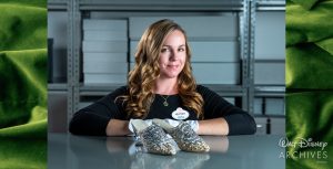 Heather Hoffman, a member of the Walt Disney Archives staff, wearing a black blouse, necklace with a green-colored pendant, and white archival gloves, stands behind a pair of gold-colored slippers, which feature iridescent silk taffeta, a plexiglass heel, silver threads and embroidered designs, and sparkling silver crystals, used in the 1998 film Ever After: A Cinderella Story. Behind her are rows of grey-colored archival boxes.