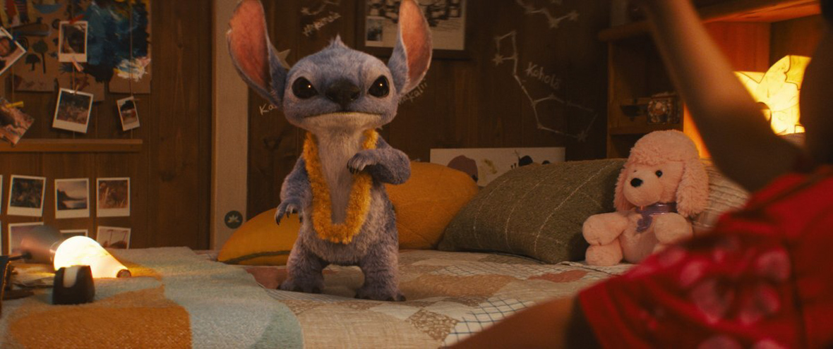 Stitch, a fuzzy blue alien with dog-like ears and features, stands in a child’s bedroom on top of the bed. He is wearing a yellow lei and staring confusedly at the camera.