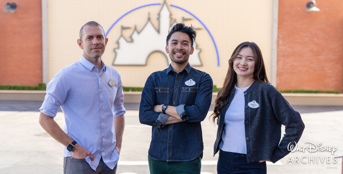 Three staff members of the Walt Disney Archives, Ty Popko (wearing a light blue shirt and wristwatch), Patrick Letrondo Markulis (wearing a dark blue shirt and watch), and Amaris Ma (wearing a light purple blouse, blue sweater, and necklace), pose outside in front of a depiction of Sleeping Beauty Castle at The Walt Disney Studios in Burbank, California.